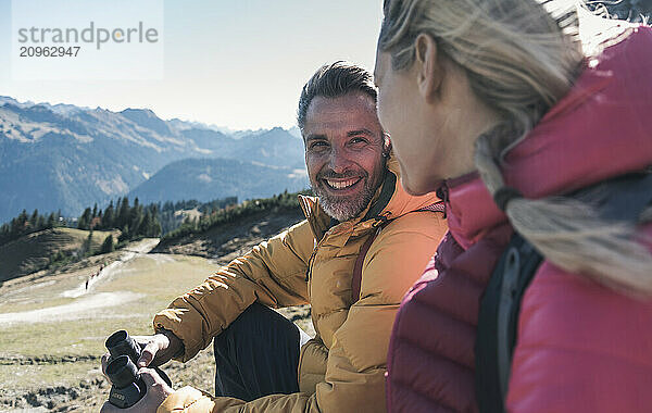 Happy man holding binocular and sitting with friend at mountain