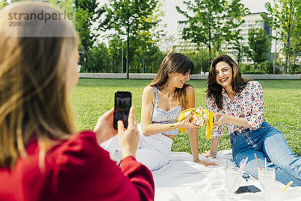 Woman photographing happy friends sitting on picnic blanket in park