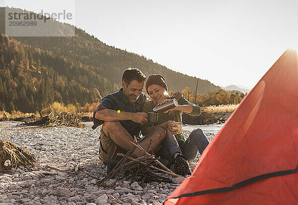 Smiling woman pouring coffee in mug and sitting with boyfriend at riverbank