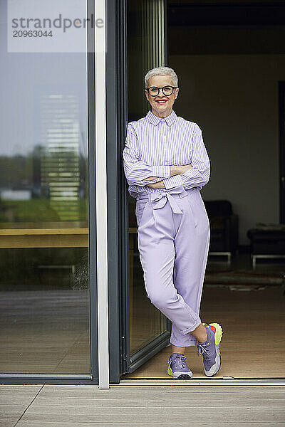 Confident senior woman leaning against balcony door at home