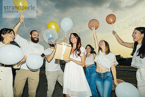 Happy friends celebrating woman's birthday with balloons and gift at sunset