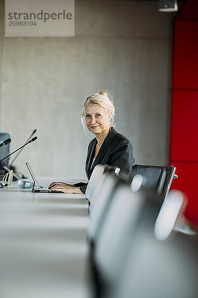 Smiling businesswoman with laptop sitting on chair in meeting room