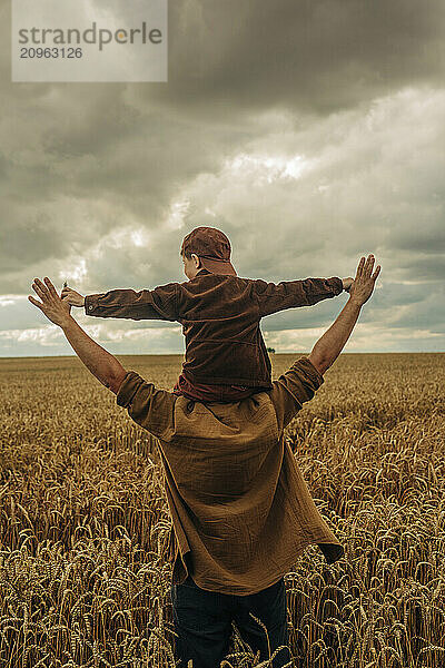 Father holding little son on shoulders in a wheat field on a cloudy day  rear view.