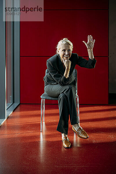 Businesswoman sitting on chair and waving near red wall at office