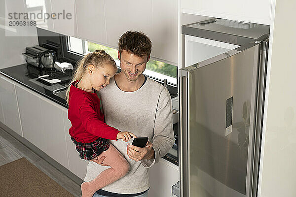 Father with daughter using smart phone near refrigerator in kitchen at home