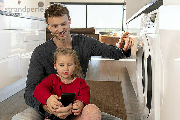 Smiling man and daughter operating washing machine through smart phone app at home