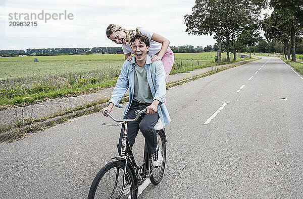 Cheerful young man riding bicycle with girlfriend on road near meadow