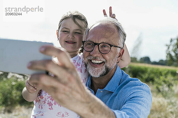 Happy mature man in eyeglasses taking selfie with granddaughter doing peace sign