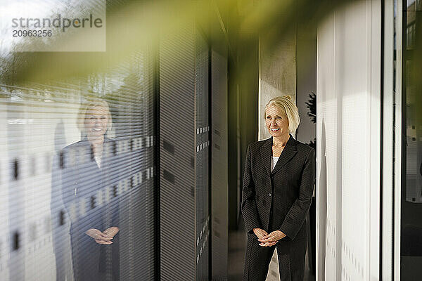 Smiling senior businesswoman standing with hands clasped at office