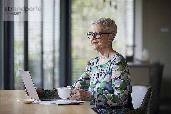 Senior woman sitting at table at home with laptop and coffee cup