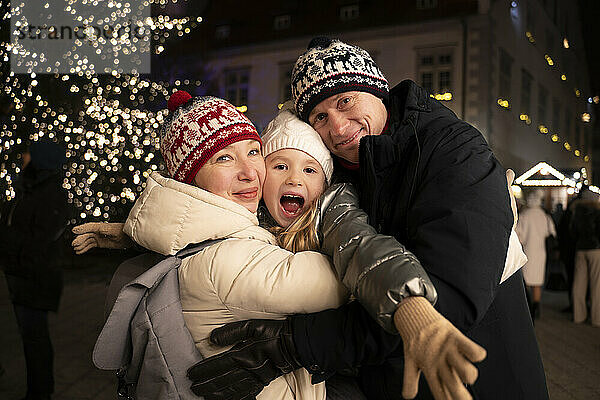 Mother and father holding daughter near christmas tree in city