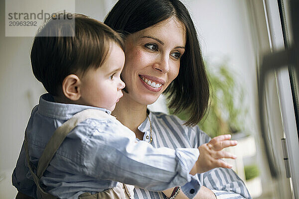 Smiling woman and baby looking through window at home