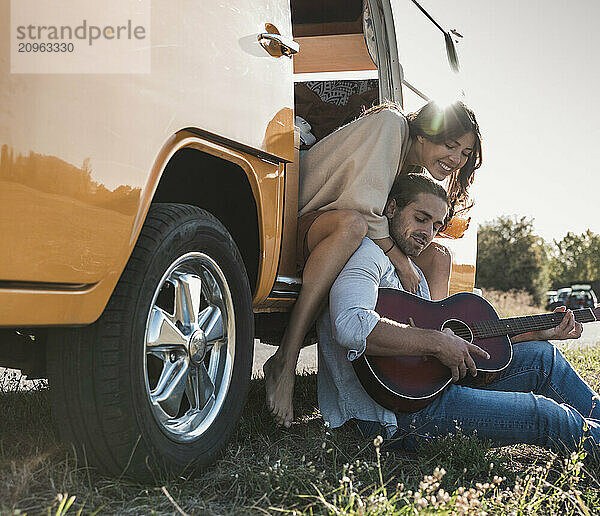 Young man playing guitar and sitting with girlfriend at doorway of camper van