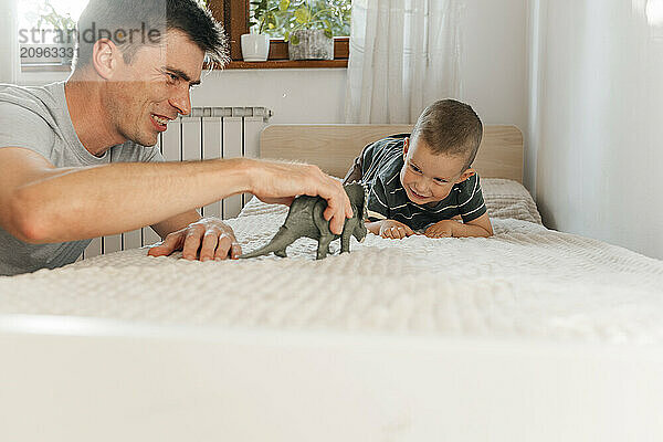 Happy father and son playing with dinosaur toy on bed at home