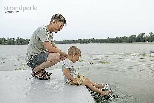 Father crouching near son sitting on pier and playing with water