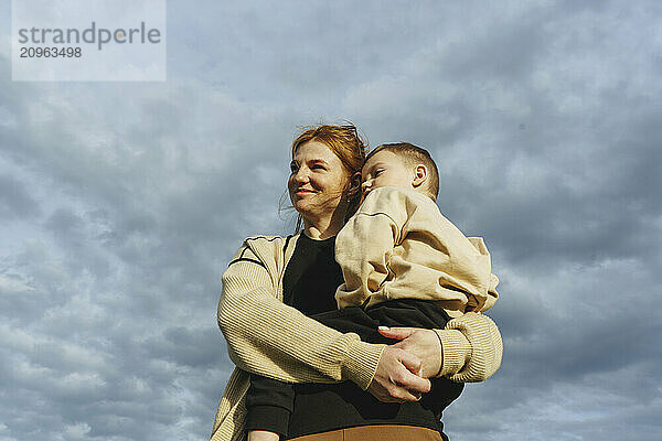 Smiling mother carrying son under cloudy sky