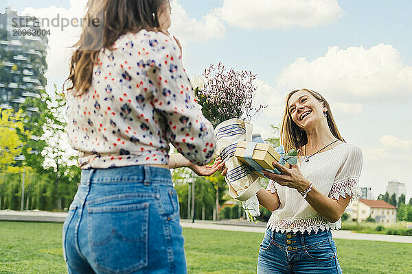 Woman giving gift box and bouquet of flowers to happy friend under sky