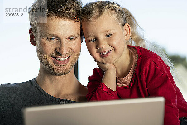 Smiling daughter leaning on elbow and watching tablet PC with father at home