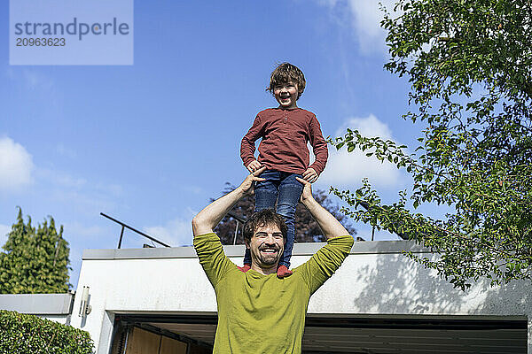 Happy boy standing on father's shoulder under blue sky