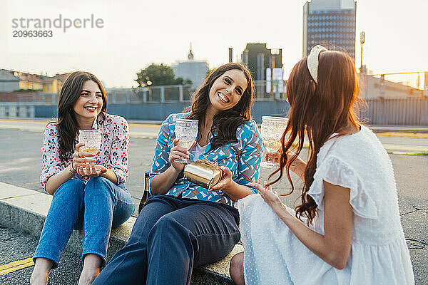 Cheerful woman with friends holding wine glasses and sitting on footpath