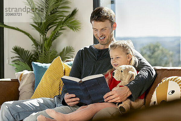 Happy father reading book with daughter sitting on sofa at home