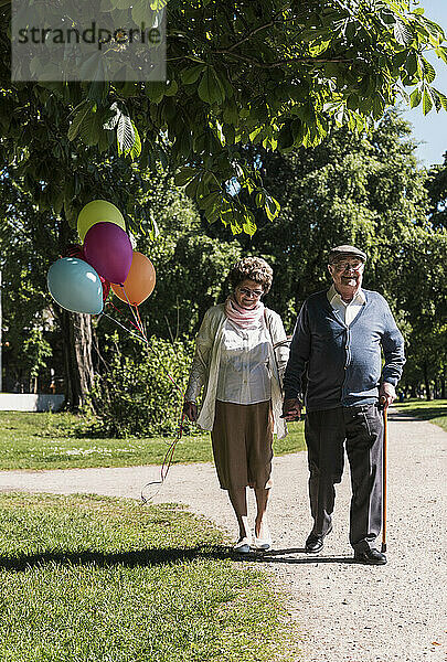 Senior couple walking with balloons in park on sunny day