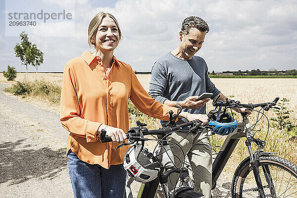 Smiling woman wheeling bicycle with man using smart phone