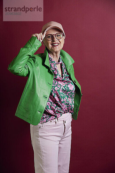 Fashionable senior woman wearing green jacket and cap against purple background