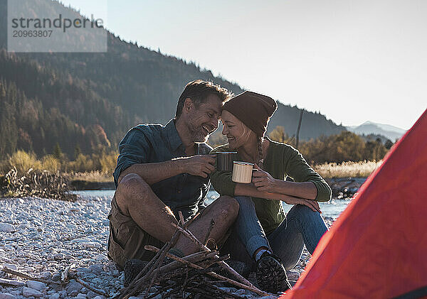 Happy man sitting with girlfriend and enjoying coffee during camping at riverside