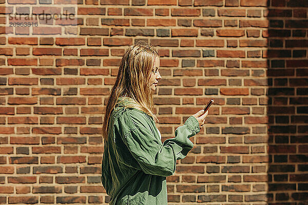 Young woman using smart phone and standing in front of brick wall