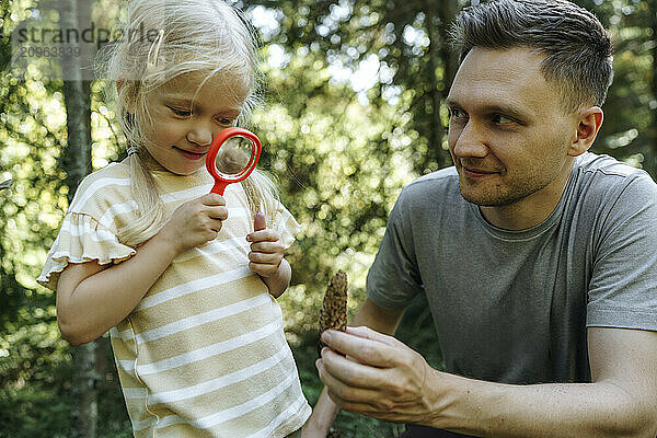 Daughter observing plant part using magnifying glass with father in forest