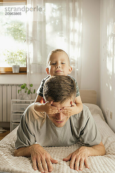 Son covering eyes of father lying on bed at home