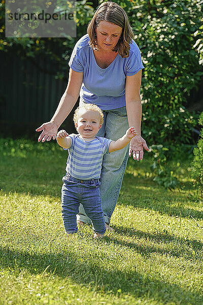 Mother supporting her son taking his first steps in a sunny garden.