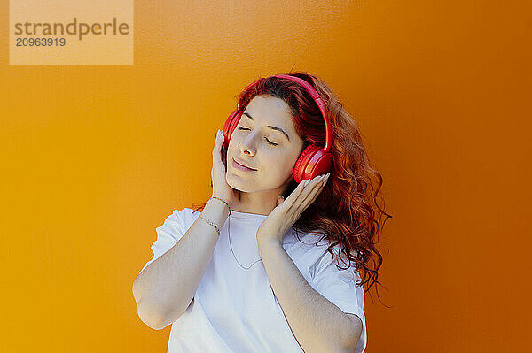 Redhead woman listening to music through wireless headphones against colored background