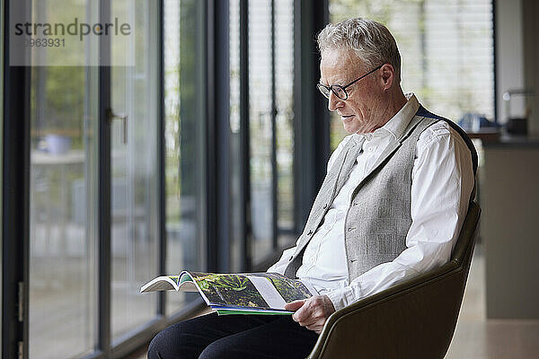 Senior man sitting in armchair at home reading magazine