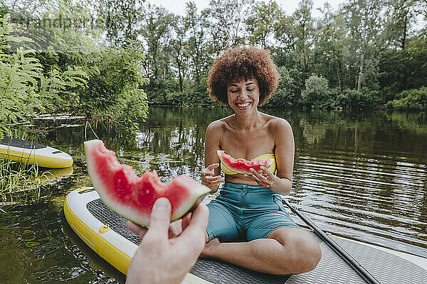 Happy woman eating watermelon with friend on paddleboard in lake