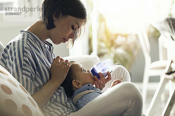 Young mother feeding baby boy with milk bottle at home