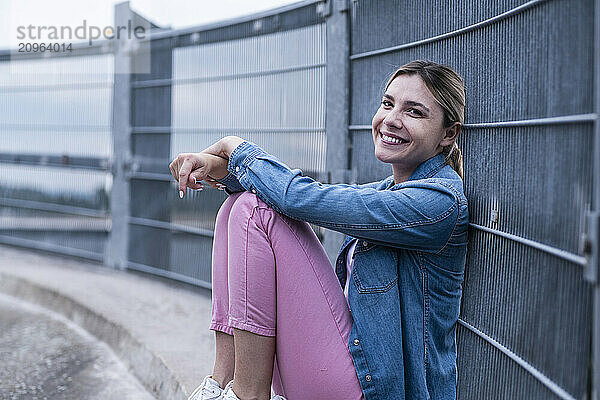 Smiling young woman with hands on knee sitting near railing