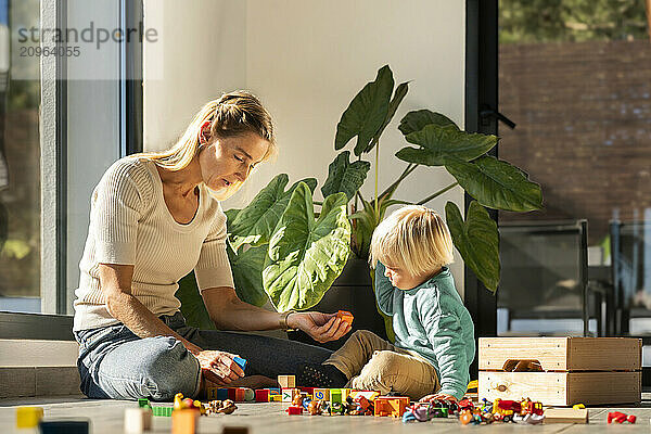 Mother assisting son playing with building blocks sitting on floor at home