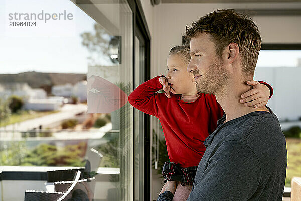 Father and daughter looking out of glass window at home