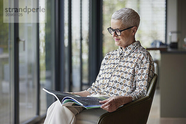 Senior woman sitting in armchair at home reading magazine