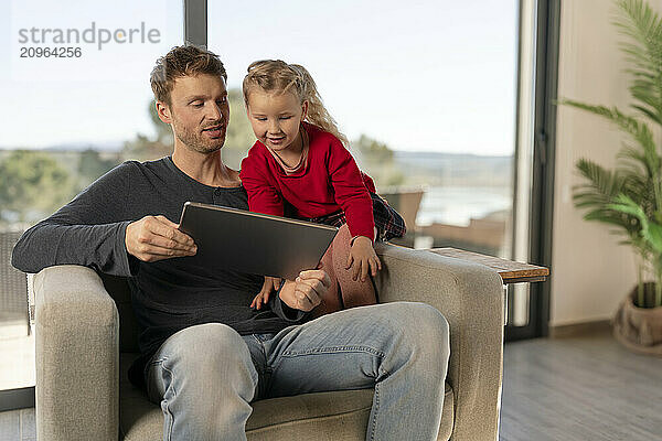 Father sitting on armchair and showing tablet PC to daughter at home