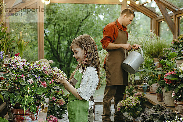 Girl looking at flowers and father watering plants in greenhouse