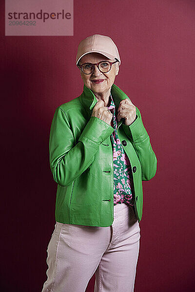 Fashionable senior woman wearing green jacket and cap against purple background