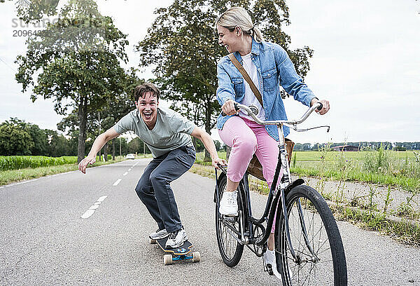 Cheerful man skateboarding with girlfriend riding bicycle on road