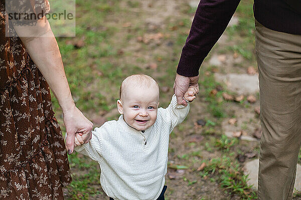 Toddler boy smiles at camera as mom and dad hold his hands.