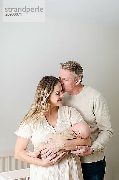 Mom holds baby while dad kisses her on the forehead.