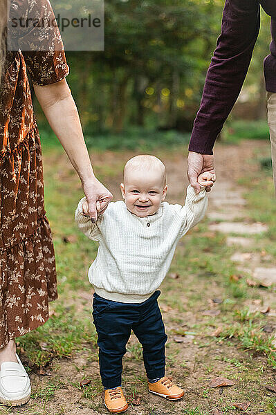 Toddler boy smiles at camera as mom and dad hold his hands.