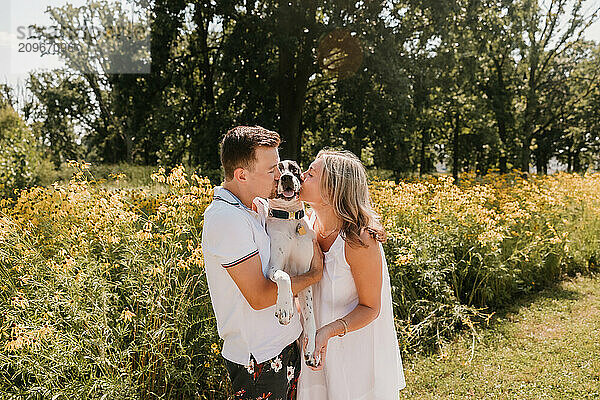 Smiling young couple kissing their dog in front of wildflowers