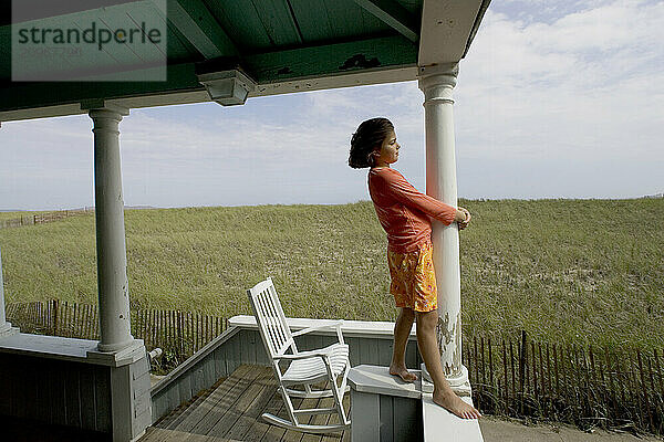 A young girl stand on the railing of a porch with sand dunes in the background.